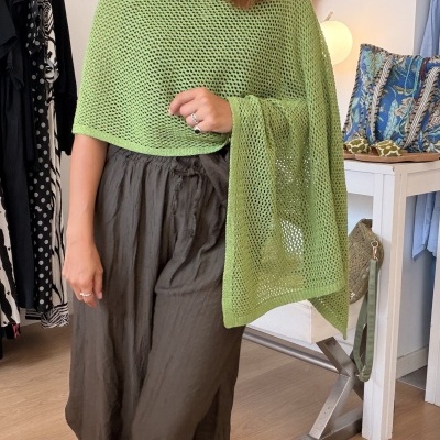 Mulher com poncho verde claro e calças castanhas em loja de roupa com decoração e acessórios.