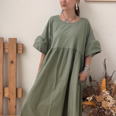 Vestido longo verde com mangas largas, bolsos e ténis bege, acessórios metálicos, em fundo com madeira, almofada e vaso com flores secas.