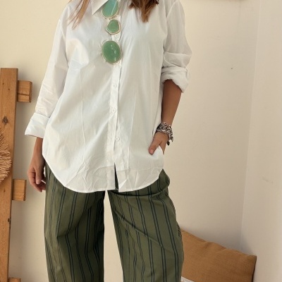 Mulher veste camisa branca, calças largas verdes com riscas, óculos de sol pendurados e pulseira, em ambiente interior minimalista