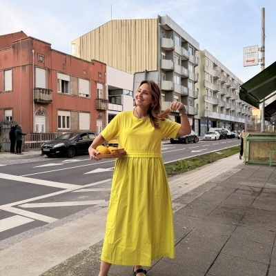 Mulher com vestido amarelo e sapatos pretos segura cesta com laranjas numa rua da cidade
