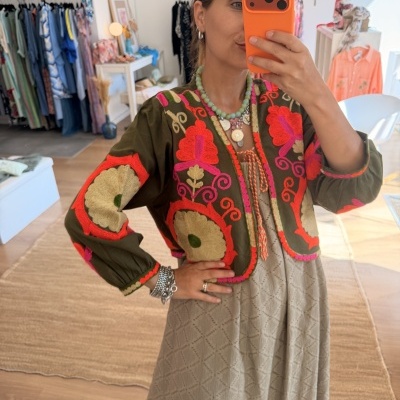 Casaco verde-oliva bordado com padrões florais vermelhos e rosa, visto em selfie numa loja de roupa