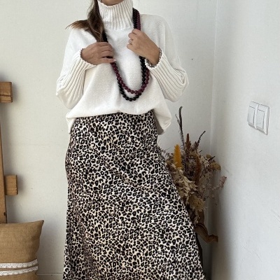 Mulher com camisola branca de gola alta, saia de leopardo e sapatilhas brancas num ambiente interior minimalista.
