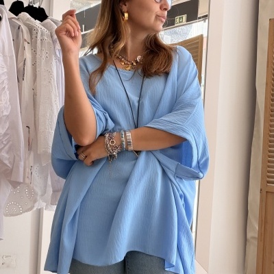 Mulher de camisola azul claro e calças de ganga com acessórios, em frente a um espelho grande