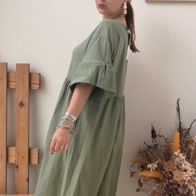Mulher com vestido verde comprido e acessórios em ambiente interior com decoração neutra.