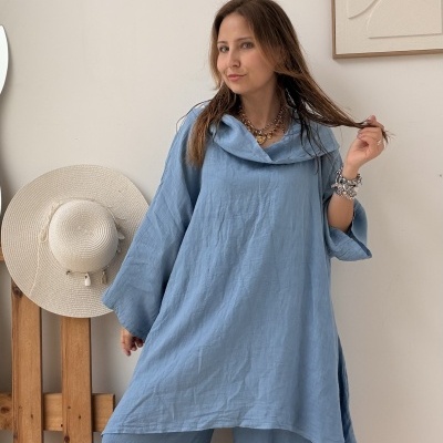 Mulher vestindo conjunto azul claro com túnica e calças largas, chapéu de palha ao fundo em estrutura de madeira
