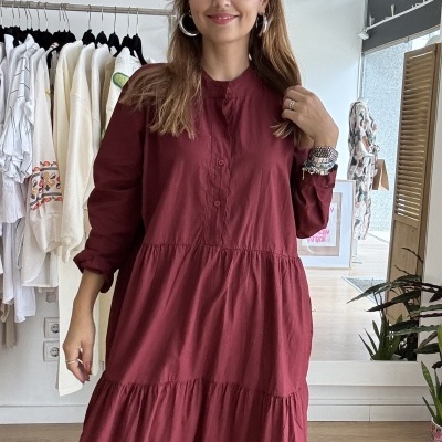 Mulher vestindo vestido bordô comprido com botões, em loja com várias roupas penduradas ao fundo