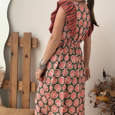 Vestido comprido com padrão de conchas cor-de-rosa sobre fundo verde, folhos às riscas nas mangas, mulher numa sala com almofada e flores secas.
