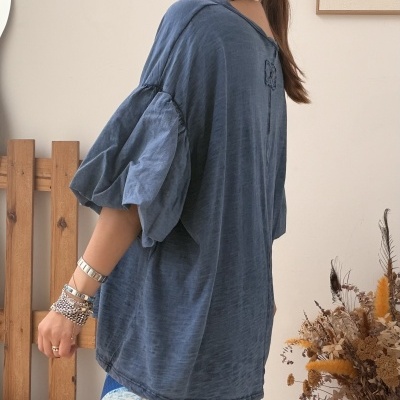 Mulher de perfil com blusa azul de mangas bufantes e saia estampada com póster e flores secas ao fundo