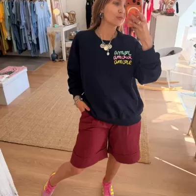 Mulher a usar sweatshirt preta com bordado colorido e bermuda borgonha, a tirar selfie em loja de roupas