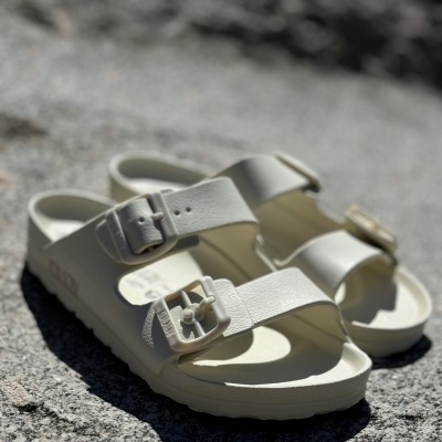 Sandálias brancas tipo Birkenstock com duas fivelas sobre superfície rochosa cinza