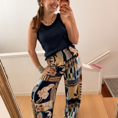 Mulher sorridente com top preto e calças largas estampadas, a tirar selfie num espelho