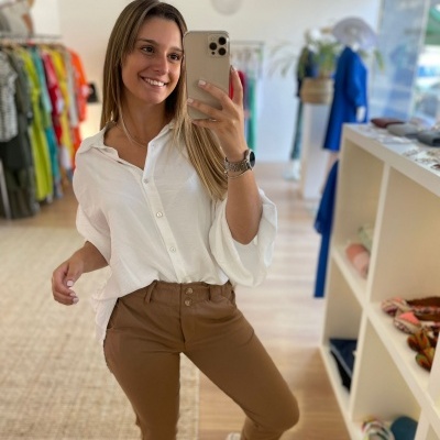 Mulher em loja de roupas com camisa branca e calças camel, a tirar selfie no espelho