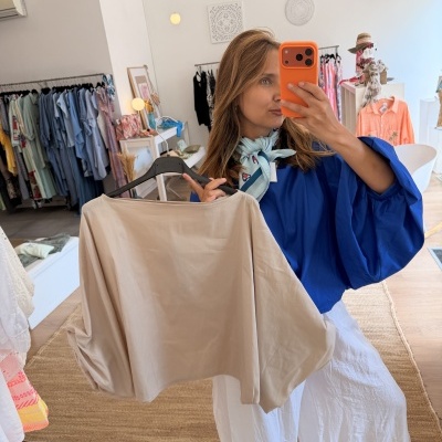 Mulher com blusa azul e lenço azul claro segurando blusa bege em cabide dentro de loja de roupa