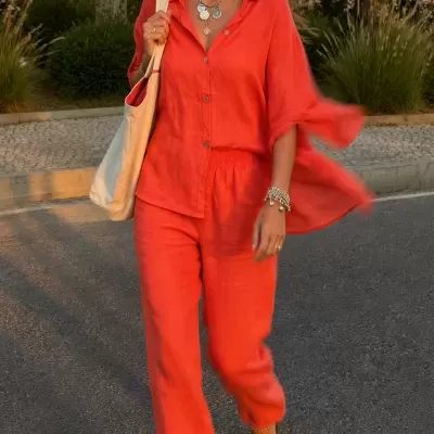 Mulher com conjunto vermelho de linho, chapéu de palha, acessórios e tote bag creme a caminhar na rua.