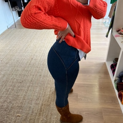 Mulher com camisola laranja e calças de ganga azul num interior de loja com chão de madeira e tapete