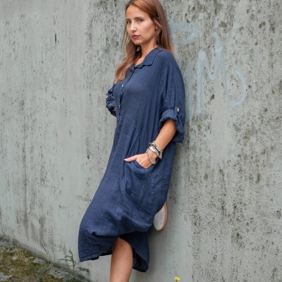 Mulher a usar vestido azul escuro comprido e sapatilhas brancas, encostada a uma parede cinzenta suja