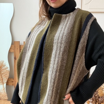 Mulher a usar colete de malha riscada verde, cinza e azul escuro sobre camisola preta de gola alta em ambiente interior.