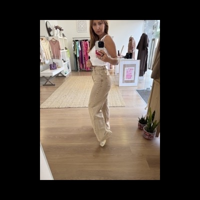 Mulher com roupa branca e calças bege largas, tirando selfie num espelho em loja de roupas.