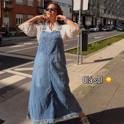 Mulher a usar vestido jardineira de ganga longo com camisa branca e ténis, a sorrir ao sol na rua