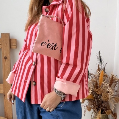 Mulher com casaco riscado rosa e vermelho, calças azuis e mala rosa com texto cest em interior decorado com flores secas