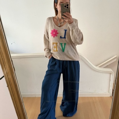 Mulher com camiseta bege com letras coloridas, calças largas azul-escuro, a tirar selfie junto a uma parede branca e chão de madeira.