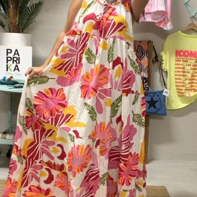 Mulher com vestido comprido floral rosa, vermelho, amarelo e verde em loja com decoração e roupas penduradas