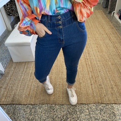 Calças de ganga azul de cintura alta com camisa colorida e ténis brancos em fundo de tapete e chão de pedra