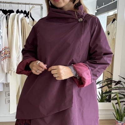 Mulher com casaco roxo de gola assimétrica e botão, com punhos rosa, num interior com roupas penduradas ao fundo