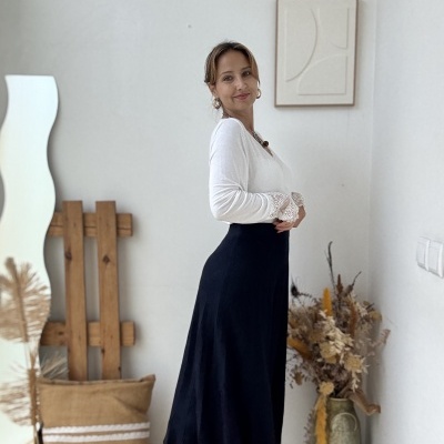 Mulher com blusa branca de manga comprida e saia preta comprida em ambiente interior decorado de forma natural.