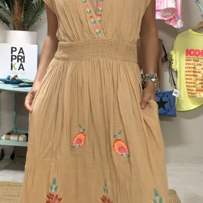 Vestido bege com bordados florais coloridos e decote em V usado por mulher em ambiente interior.