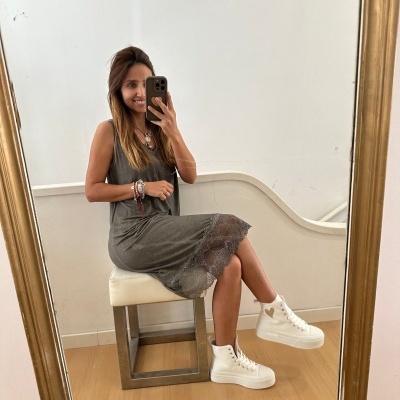 Mulher sentada num banco usando vestido cinzento com renda e sapatilhas brancas, refletida num espelho com moldura dourada.