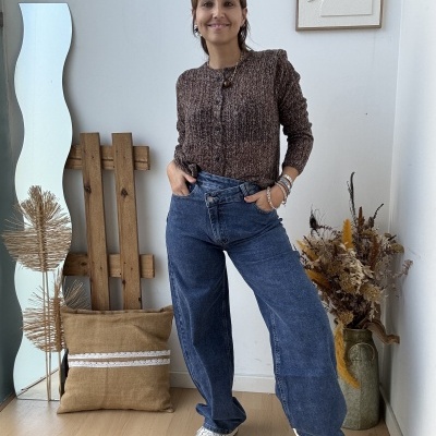 Mulher com cardigan castanho e jeans azuis em ambiente interior minimalista.