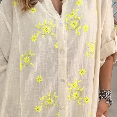 Camisa bege bordada com flores amarelas e verdes, botões na frente e mangas dobradas.
