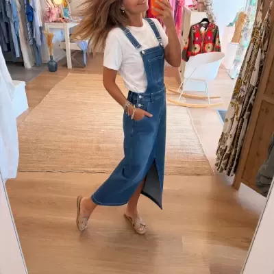 Mulher a tirar selfie com vestido azul de ganga e t-shirt branca, numa loja de roupa com várias peças coloridas ao fundo.