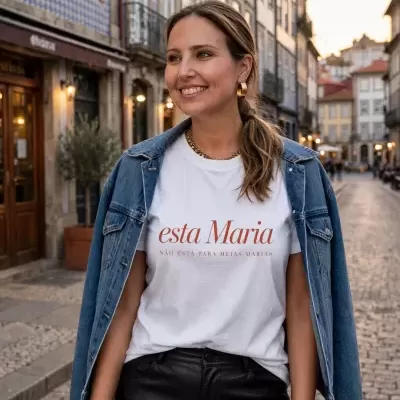 Mulher com t-shirt branca com texto, casaco de ganga azul claro e calças pretas numa rua de pedra