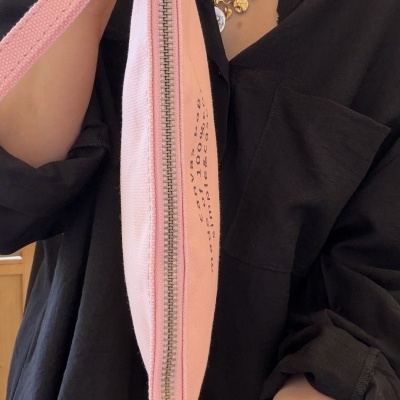 Carteira rosa de tecido com fecho metálico e texto impresso sendo segurada por pessoa com camisa preta e acessórios prateados.