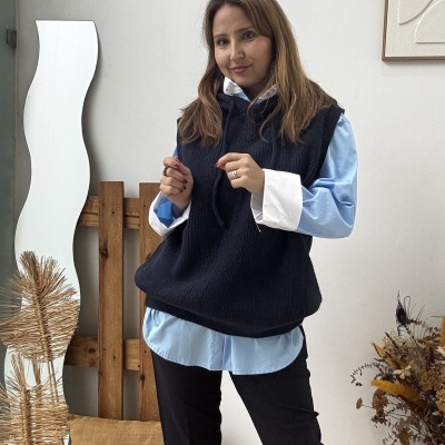 Mulher com colete preto e camisa azul num ambiente interior com espelho ondulado e arranjo de plantas secas.