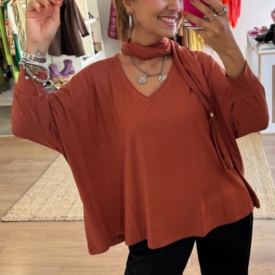 Pessoa a tirar selfie com blusa castanho avermelhado e calças pretas num espaço com roupas penduradas