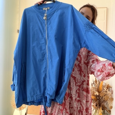 Casaco azul claro com fecho de correr e bolsos, sendo segurado por uma mulher com vestido vermelho e branco.