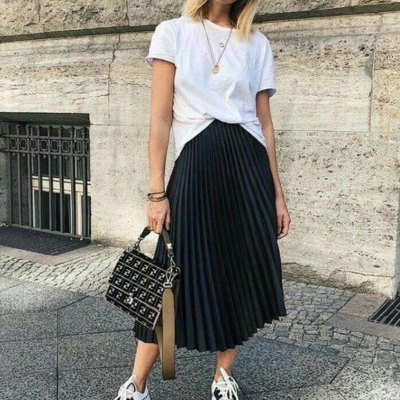 Mulher com t-shirt branca, saia plissada preta e sapatilhas com mala preta na rua