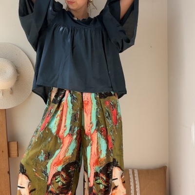 Pessoa com blusa azul escura de manga larga e calças largas coloridas com padrão artístico, parede branca e chapéu pendurado