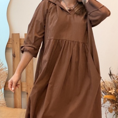 Vestido castanho comprido com botas bege e acessórios metálicos em ambiente interior