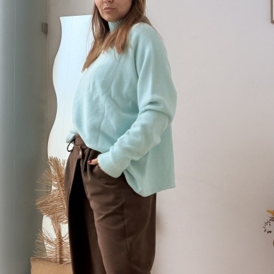 Pessoa com camisola azul clara e calças castanhas em interior com parede branca.