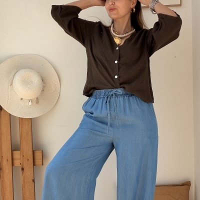 Mulher com camisa castanha, calças azuis e sapatilhas rosas junto a parede branca com chapéu de palha e almofada