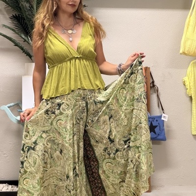 Mulher com top verde e saia comprida estampada segura a saia, cenário com planta, prateleira azul e decoração amarela