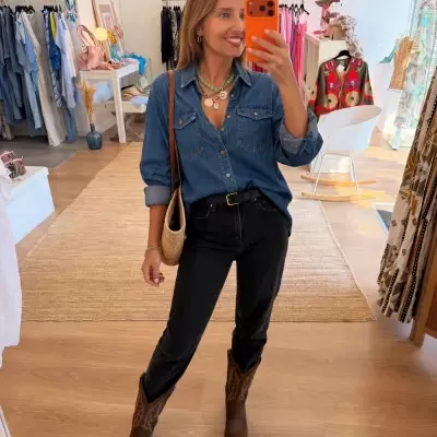 Mulher com camisa de ganga azul, calças pretas, botas castanhas e telemóvel com capa laranja a tirar selfie em loja de roupa.