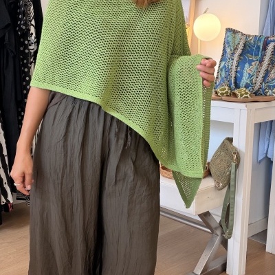 Pessoa com poncho verde e calças cáqui em quarto com decoração e roupa pendurada.