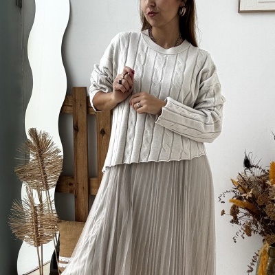 Mulher com camisola de malha branca e saia plissada bege, botas castanhas, em ambiente interior com espelho, decoração e parede branca.