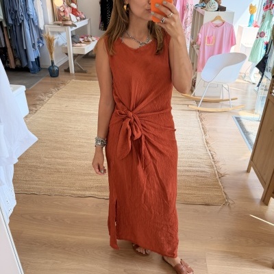Mulher a tirar selfie num espelho de corpo inteiro, vestindo vestido terracota com laço à frente e sandálias castanhas, em loja de roupa