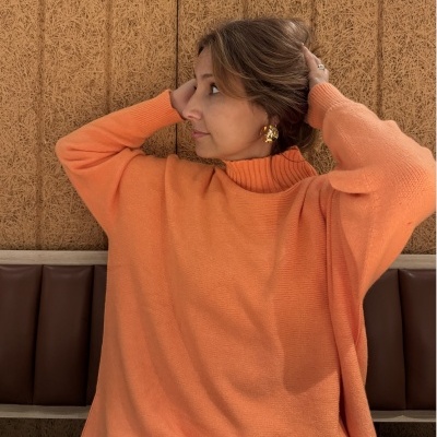 Camisola laranja de malha com gola subida, mulher sentada com brincos dourados e fundo de cortiça